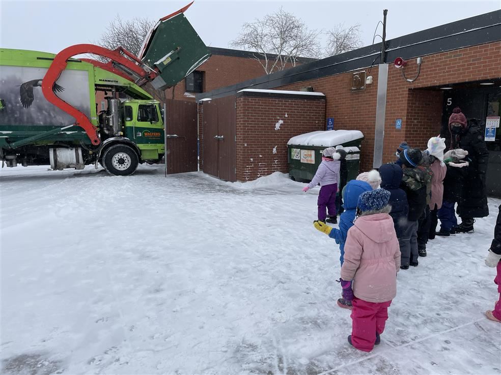 Children in snowsuits on a snowy sidewalk by a school watch a recycling truck at work.